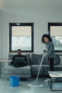 A man works on a laptop while a woman mops the floor in a cozy home setting.